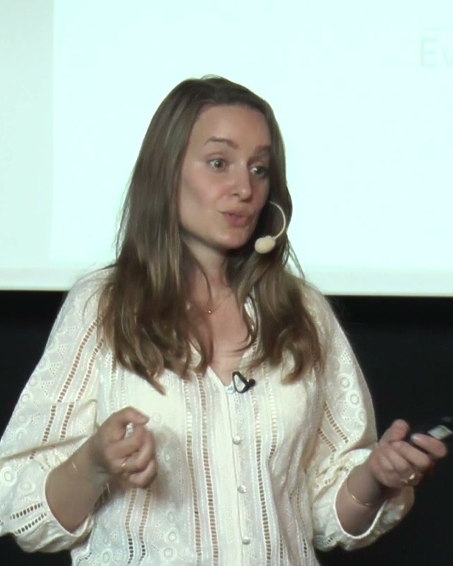 Une conférencière au micro-casque, souriante, donne sa conférence sur scène.