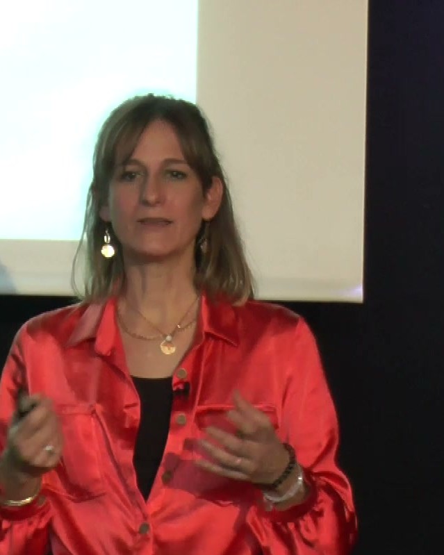 Une conférencière en chemisier rouge, micro-cravate, s'exprime en direct sur scène.