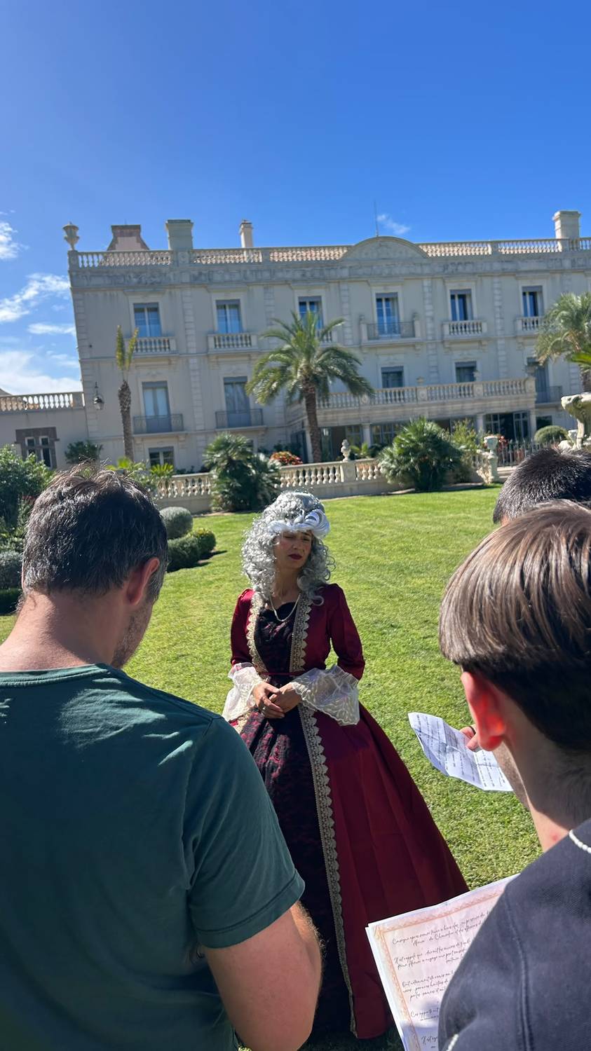 La reine de la murder party MEEUS, robe bordeaux et perruque poudree, dans les jardins du Chateau l'Hermitage de Combas, avec deux participants de dos consultant leurs indices