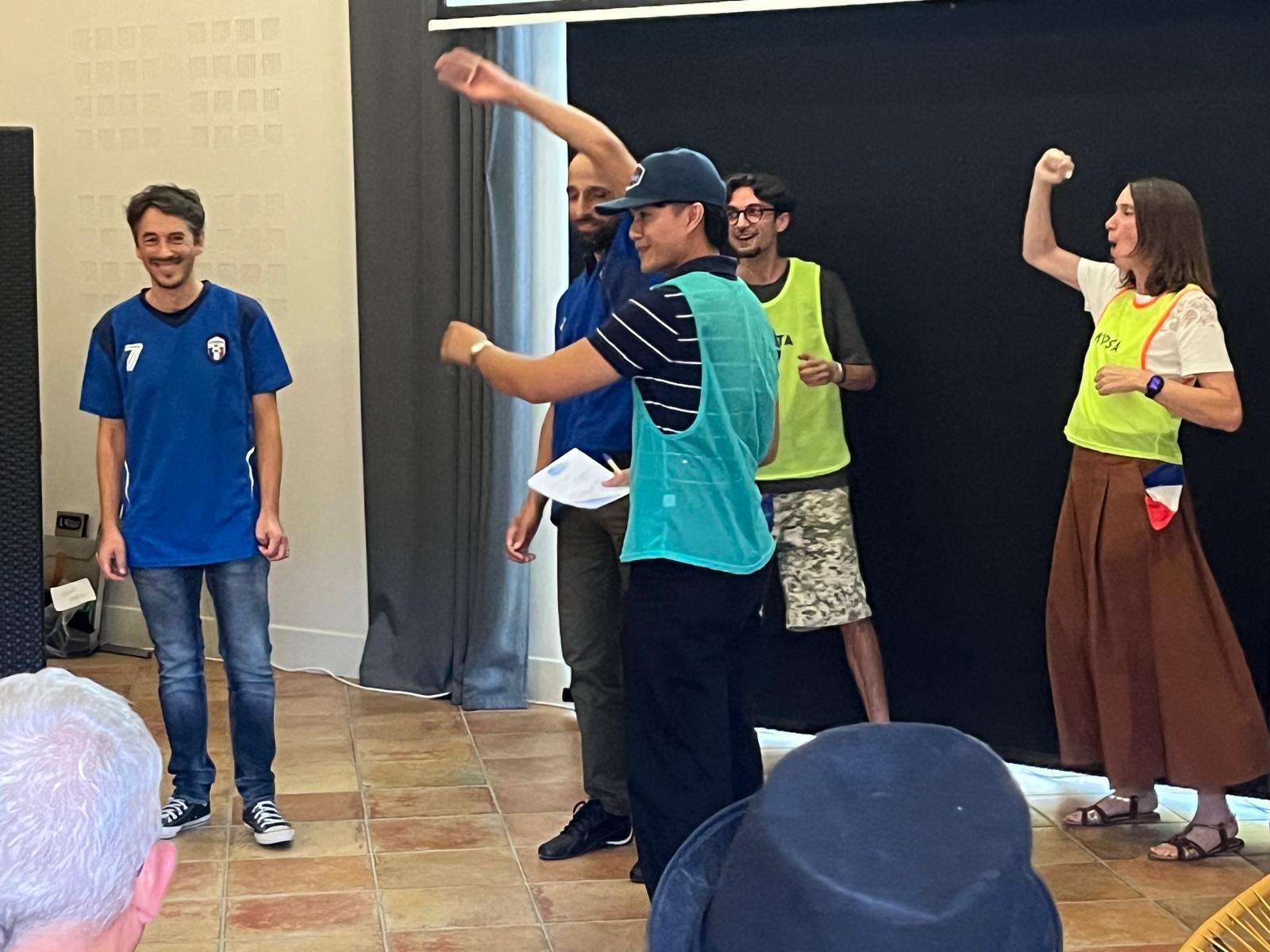 Sur le plateau, une équipe en maillots et chasubles joue une séquence sportive devant le public assis