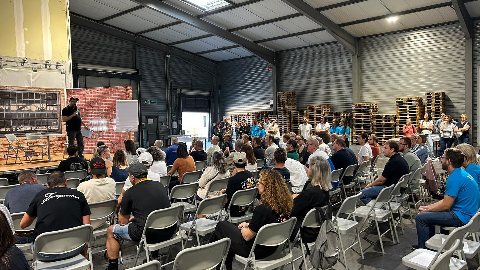 Public assis dans un hangar logistique pendant un theatre forum, un comedien de MEEUS sur une estrade avec un micro face a la salle