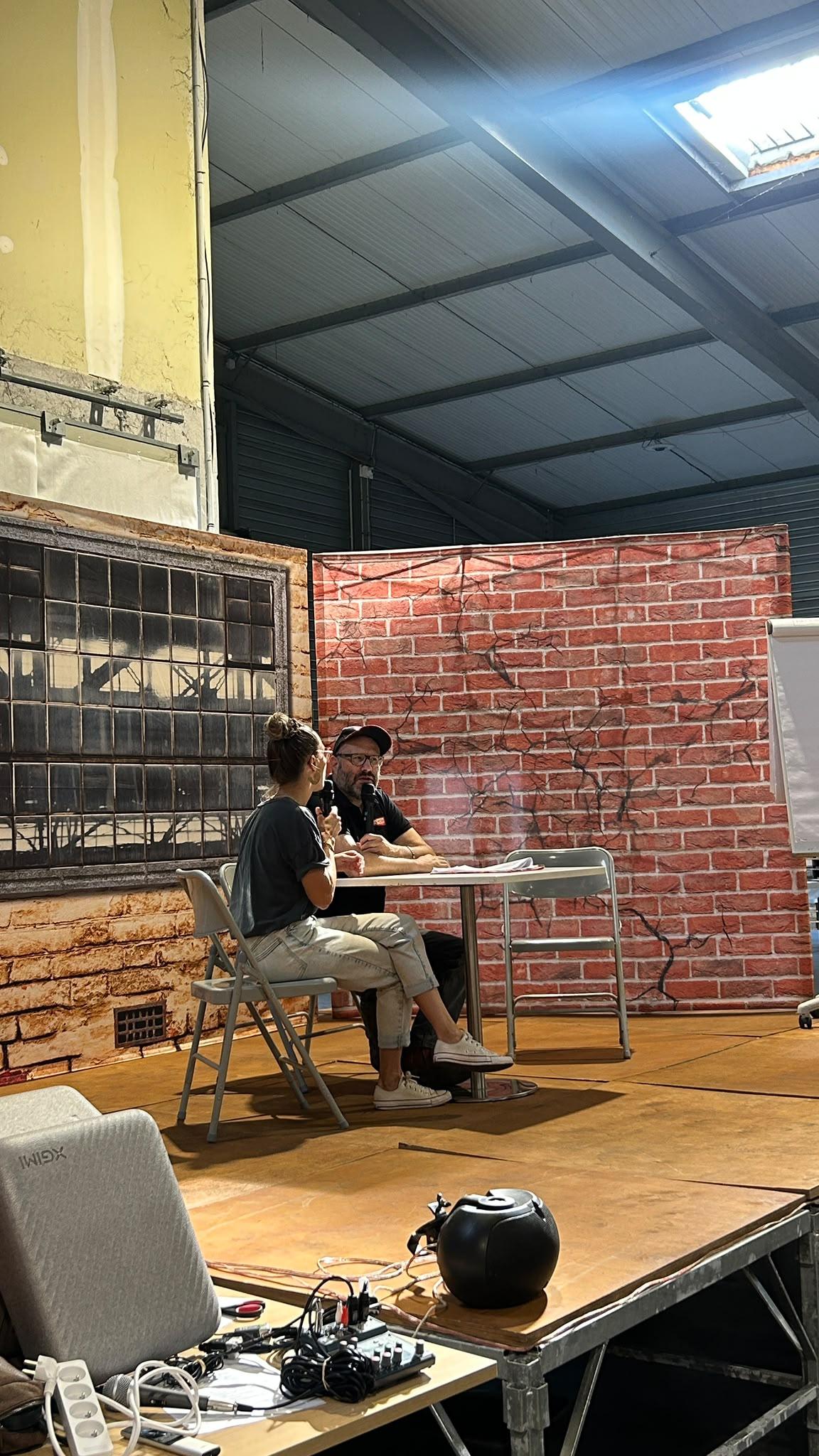 Deux comédiens de MEEUS assis à une table sur scène, micros en main, devant le décor de mur de brique peint